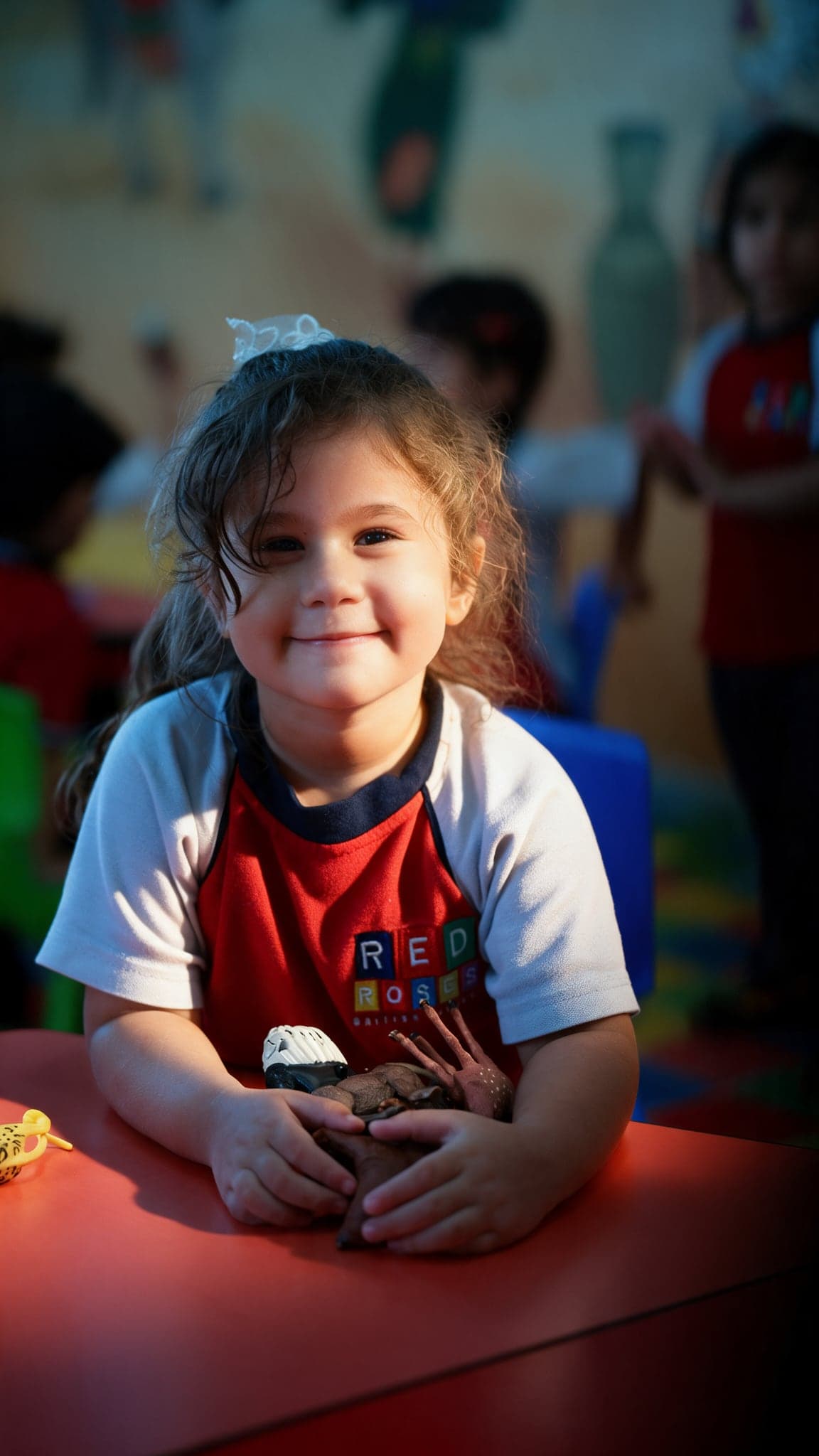 A smiling girl in the Red Roses uniform holding toy animals at a red classroom table