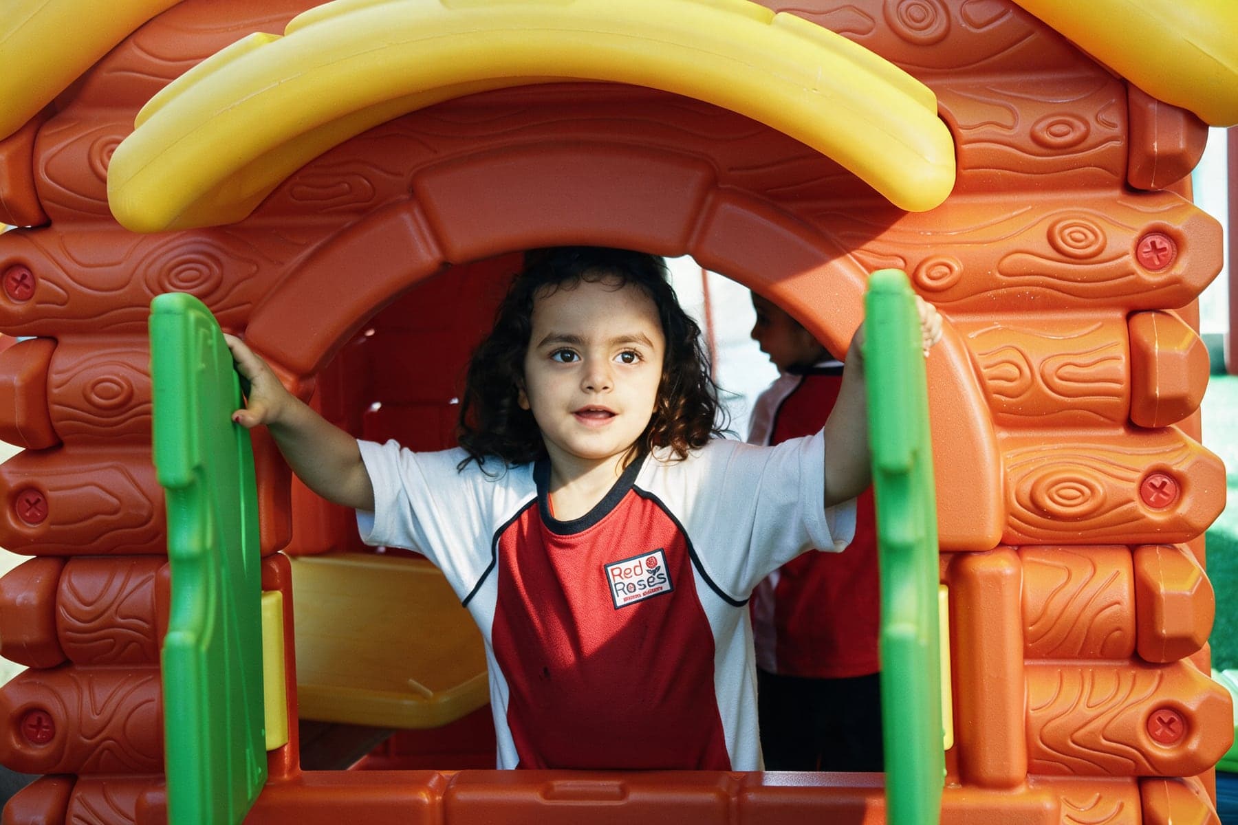 A child in the Red Roses sports uniform looking out of the window of an orange and green toy playhouse