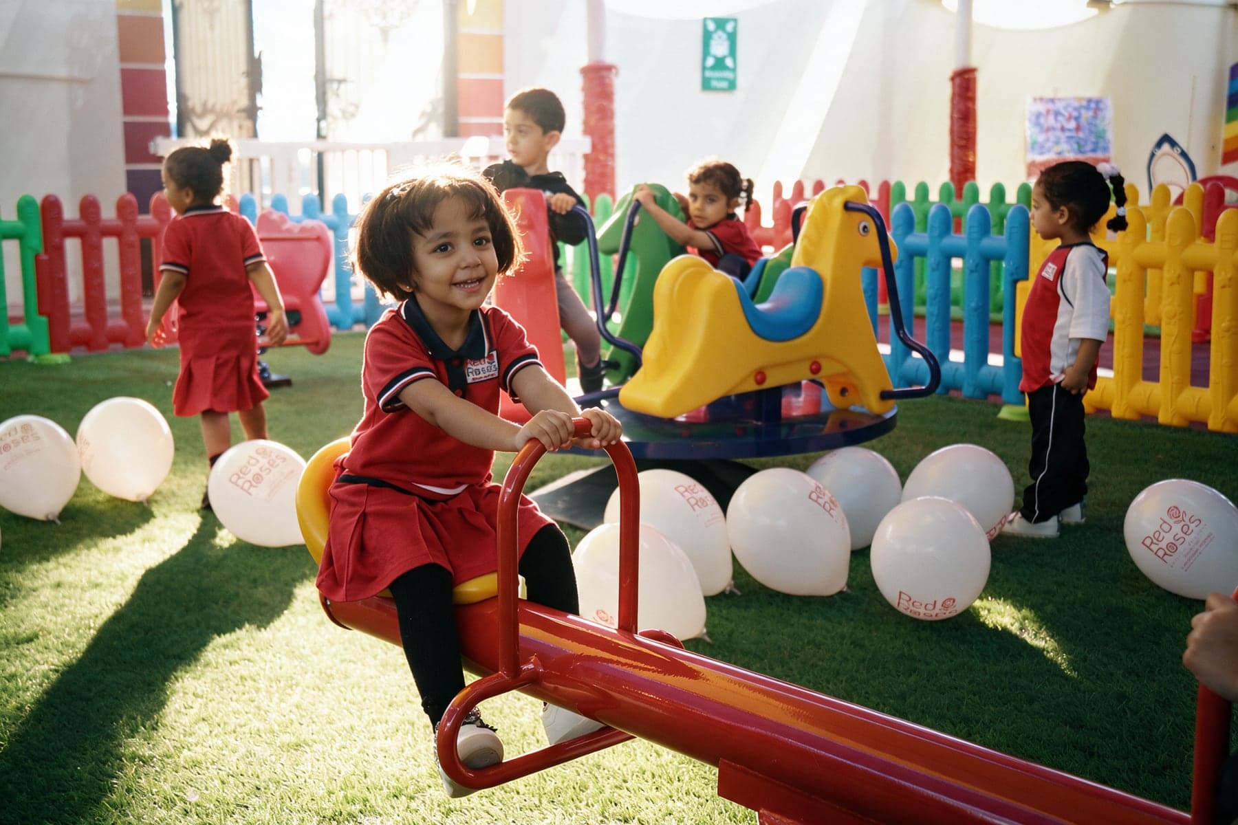 A smiling girl in the Red Roses uniform on a red seesaw in the play area, with classmates, coloured fences and Red Roses balloons behind her